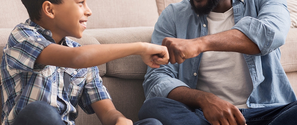 A smiling father and son sitting on the living room floor giving a fist bump.